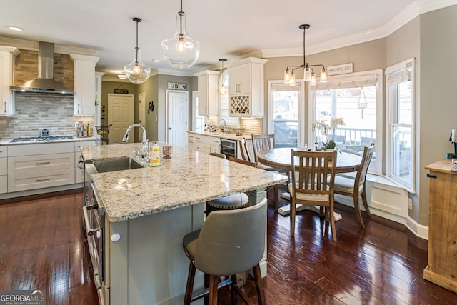 kitchen featuring dark wood-style flooring, ornamental molding, white cabinets, a sink, and wall chimney range hood