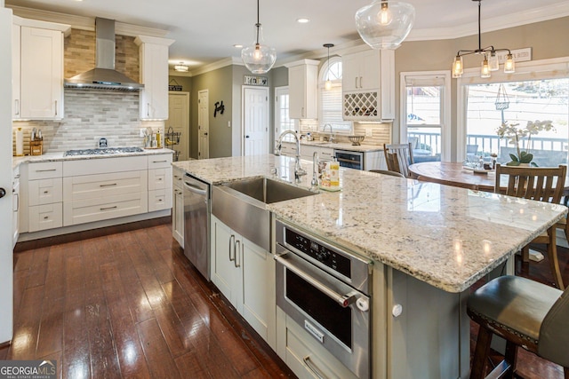 kitchen featuring a center island with sink, decorative backsplash, ornamental molding, stainless steel appliances, and wall chimney range hood