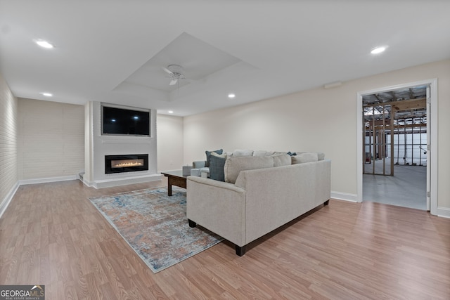 living room featuring a ceiling fan, baseboards, light wood finished floors, a tray ceiling, and a glass covered fireplace