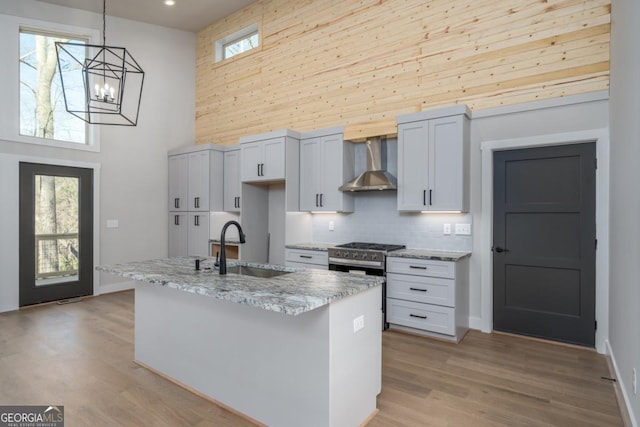 kitchen with a sink, a towering ceiling, wall chimney range hood, light wood finished floors, and gas stove