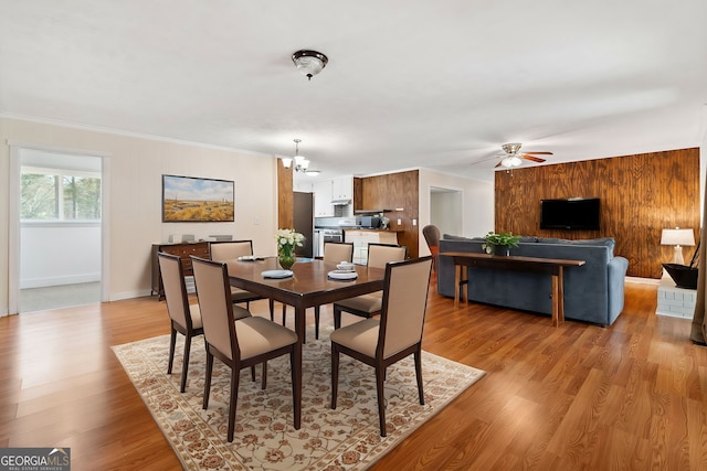 dining area with wooden walls, baseboards, crown molding, light wood-style floors, and ceiling fan with notable chandelier