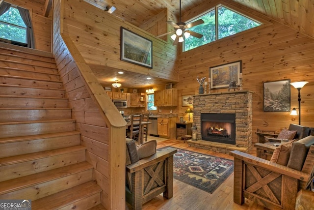 living room featuring high vaulted ceiling, a stone fireplace, wooden walls, stairs, and light wood-type flooring