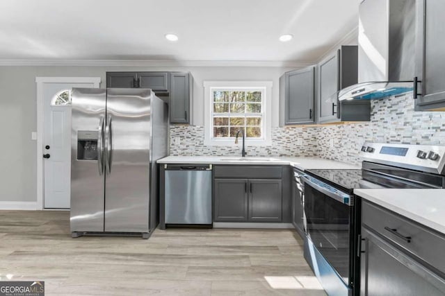 kitchen featuring appliances with stainless steel finishes, crown molding, gray cabinetry, wall chimney range hood, and a sink