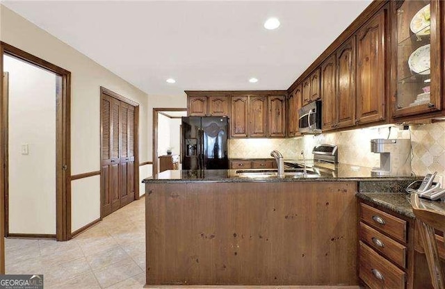 kitchen with decorative backsplash, dark stone counters, appliances with stainless steel finishes, glass insert cabinets, and a peninsula