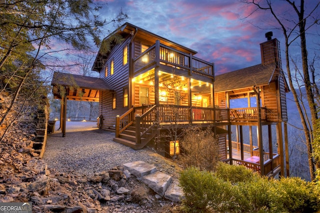 back of house at dusk featuring a balcony, roof with shingles, a chimney, and log veneer siding