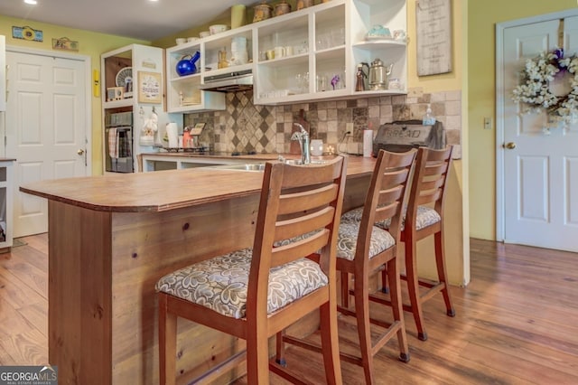 bar featuring backsplash, wood finished floors, oven, under cabinet range hood, and a sink