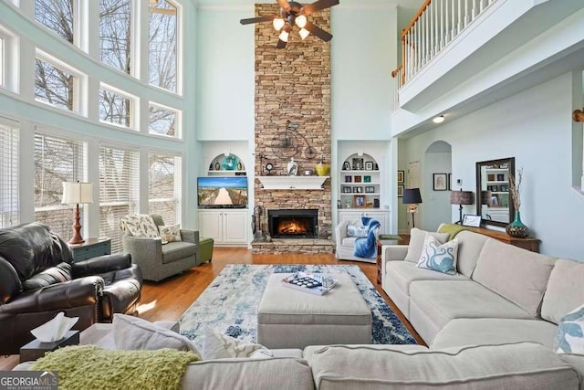 living area featuring arched walkways, ceiling fan, a stone fireplace, built in shelves, and wood finished floors