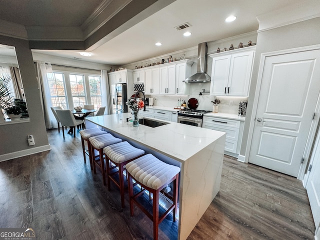 kitchen with visible vents, appliances with stainless steel finishes, a kitchen island with sink, a sink, and wall chimney exhaust hood