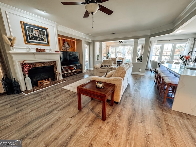 living area featuring light wood-style flooring, a fireplace, a ceiling fan, ornamental molding, and decorative columns