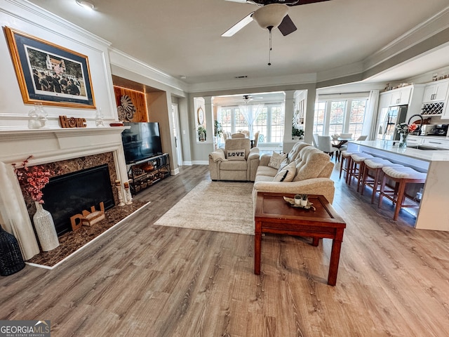 living area with a fireplace, a ceiling fan, light wood-type flooring, decorative columns, and crown molding
