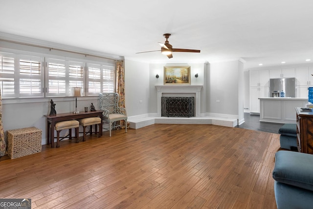 living room with dark wood-style flooring, crown molding, a fireplace with raised hearth, ceiling fan, and baseboards