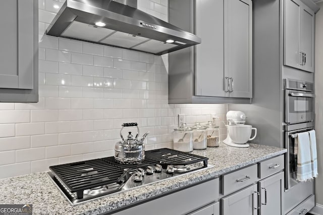 kitchen with light stone counters, decorative backsplash, gray cabinetry, appliances with stainless steel finishes, and wall chimney range hood