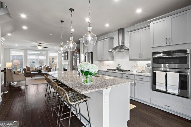 kitchen featuring a center island with sink, a breakfast bar area, stainless steel appliances, dark wood-type flooring, and wall chimney exhaust hood