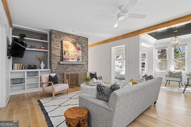 living area with built in features, beam ceiling, a stone fireplace, wood finished floors, and ceiling fan with notable chandelier