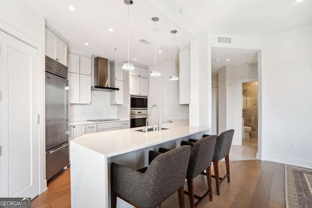 kitchen featuring visible vents, a sink, wall chimney range hood, built in appliances, and a peninsula