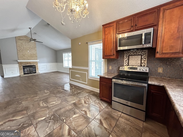 kitchen featuring lofted ceiling, a wainscoted wall, appliances with stainless steel finishes, a stone fireplace, and ceiling fan with notable chandelier