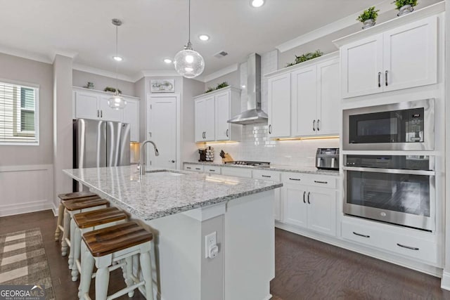 kitchen featuring stainless steel appliances, a sink, wall chimney exhaust hood, a center island with sink, and crown molding