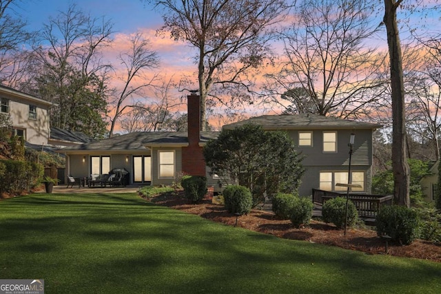 back of house at dusk with brick siding, a patio area, a chimney, and a yard