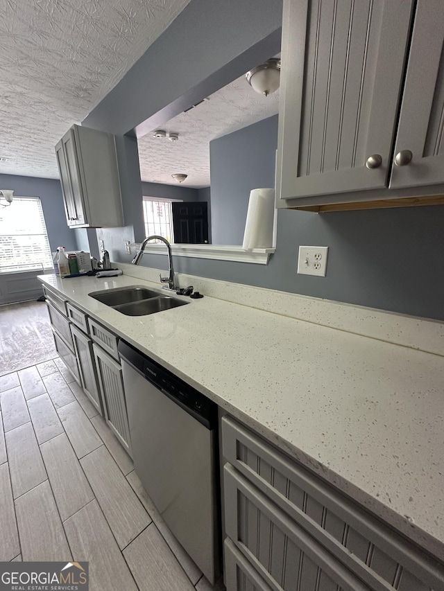 kitchen with a textured ceiling, light stone counters, gray cabinetry, a sink, and stainless steel dishwasher