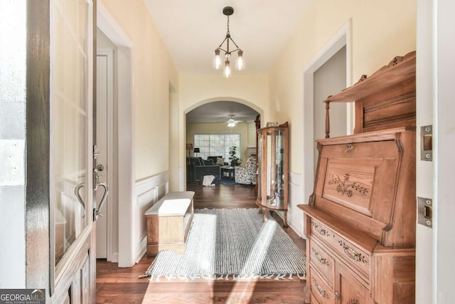 corridor featuring arched walkways, a wainscoted wall, dark wood finished floors, a notable chandelier, and a decorative wall