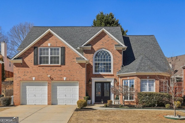 traditional-style house with french doors, brick siding, roof with shingles, an attached garage, and driveway