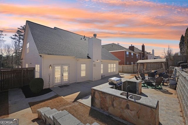 back of house at dusk featuring a fenced backyard, exterior kitchen, french doors, a chimney, and a patio area