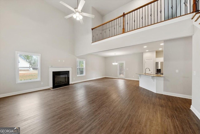 unfurnished living room featuring dark wood-style flooring, a glass covered fireplace, a sink, baseboards, and ceiling fan with notable chandelier