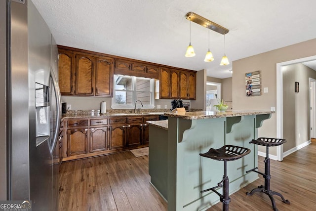 kitchen featuring a breakfast bar area, a sink, hanging light fixtures, stainless steel refrigerator with ice dispenser, and dark wood-style floors