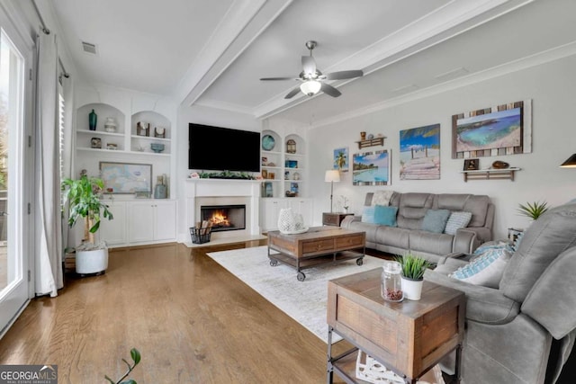 living room with built in features, a ceiling fan, a fireplace with flush hearth, beamed ceiling, and wood finished floors