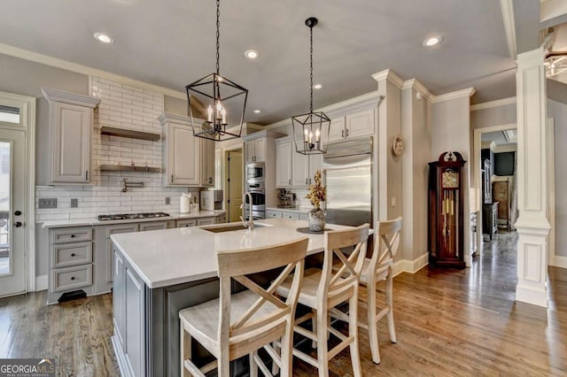 kitchen featuring decorative columns, an island with sink, built in appliances, light countertops, and a sink