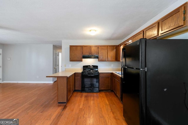 kitchen featuring under cabinet range hood, a peninsula, light countertops, brown cabinets, and black appliances