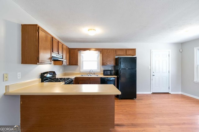 kitchen featuring brown cabinets, a peninsula, under cabinet range hood, light countertops, and black appliances