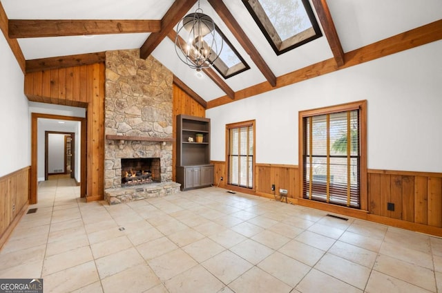 unfurnished living room with light tile patterned floors, a wainscoted wall, beamed ceiling, a stone fireplace, and a notable chandelier