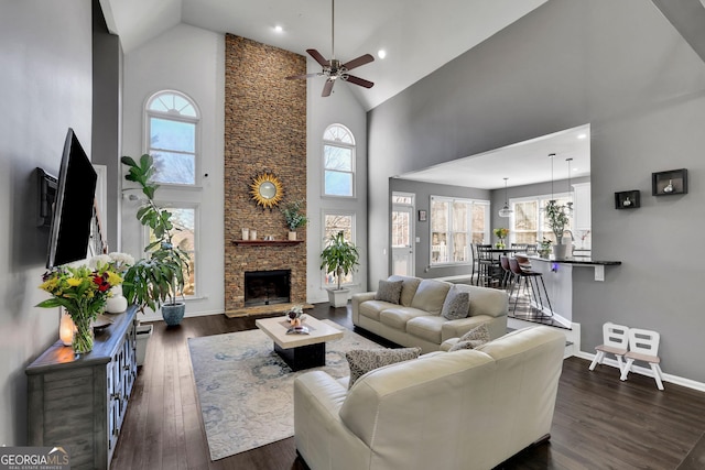 living room with baseboards, a ceiling fan, dark wood-style floors, a stone fireplace, and high vaulted ceiling