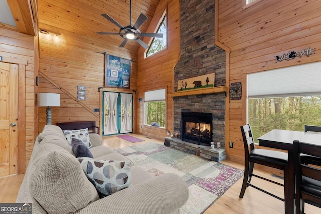 living area with wood finished floors, a stone fireplace, wooden walls, high vaulted ceiling, and a ceiling fan