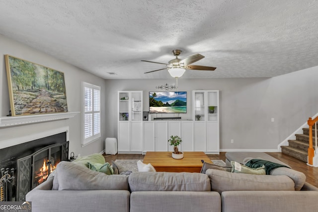 living area featuring a ceiling fan, wood finished floors, a lit fireplace, stairs, and a textured ceiling