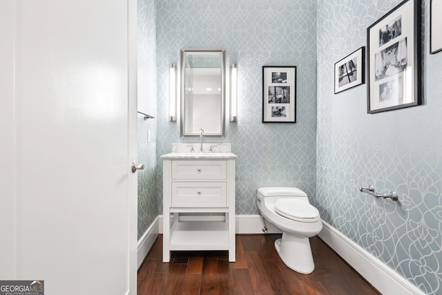 half bathroom with wallpapered walls, baseboards, toilet, wood finished floors, and vanity