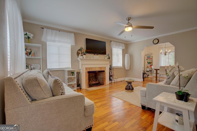 living area with light wood-style flooring, ceiling fan with notable chandelier, baseboards, a tiled fireplace, and crown molding