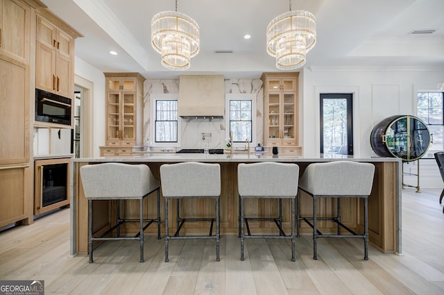 kitchen with an inviting chandelier, custom exhaust hood, a tray ceiling, light brown cabinetry, and light wood-type flooring