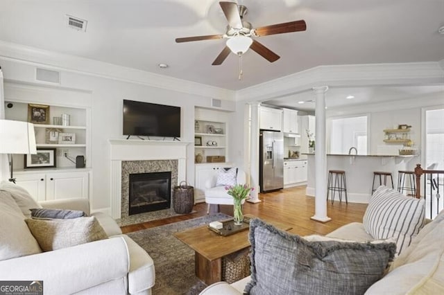 living area featuring built in shelves, a fireplace with flush hearth, light wood-style floors, decorative columns, and crown molding