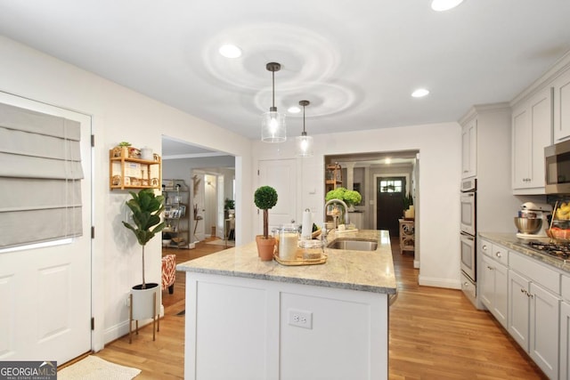 kitchen with light stone counters, a sink, hanging light fixtures, light wood-type flooring, and an island with sink