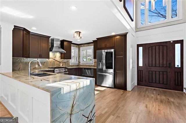 kitchen with light stone counters, stainless steel appliances, dark brown cabinets, a peninsula, and wall chimney exhaust hood