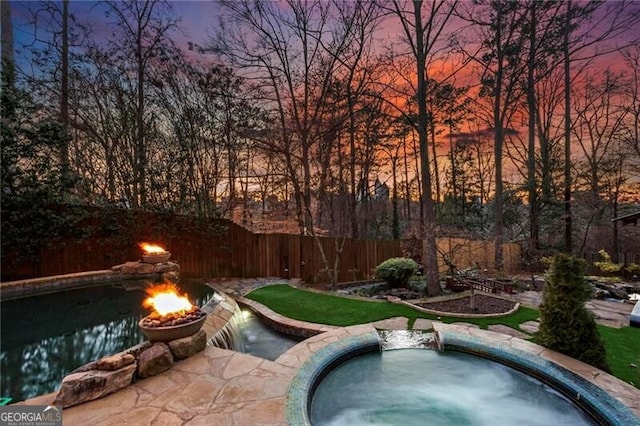 pool at dusk featuring an outdoor fire pit, fence, and an in ground hot tub