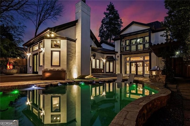 back of house at dusk featuring an outdoor pool, a balcony, a chimney, french doors, and stucco siding