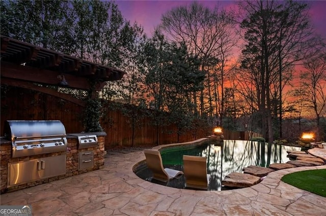 patio terrace at dusk with grilling area, an outdoor kitchen, fence, and a fenced in pool