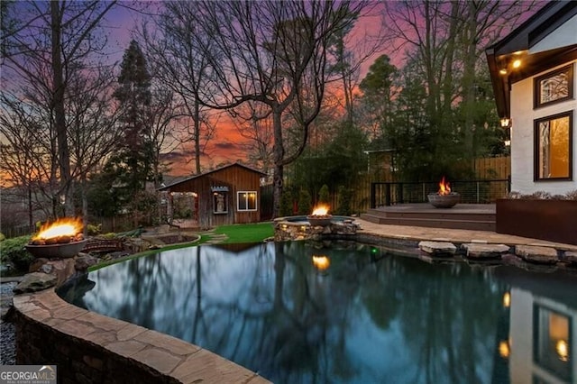 pool at dusk featuring a fire pit, an outdoor pool, a storage structure, fence, and an outdoor structure