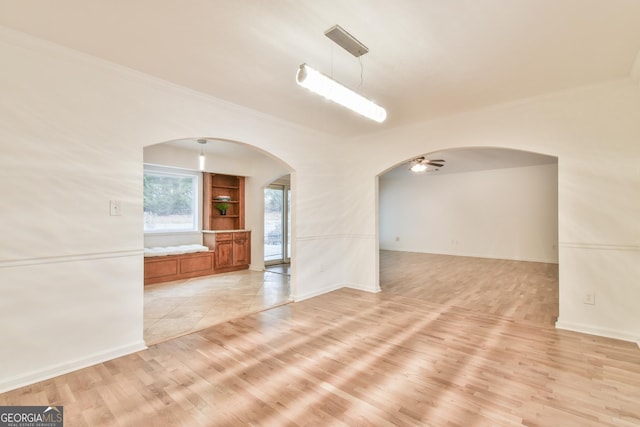 spare room featuring a ceiling fan, arched walkways, light wood-style floors, crown molding, and baseboards