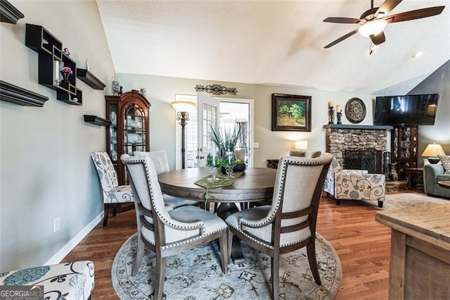 dining area featuring baseboards, lofted ceiling, ceiling fan, wood finished floors, and a stone fireplace