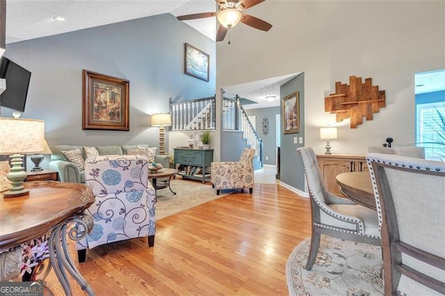 living area featuring baseboards, a ceiling fan, light wood-style flooring, stairway, and high vaulted ceiling