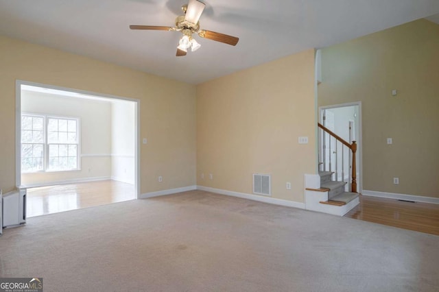 carpeted empty room featuring a ceiling fan, visible vents, baseboards, and stairs